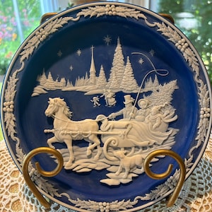 May include: A blue decorative plate featuring a white relief scene of a horse-drawn sleigh in a winter landscape. The plate has a white floral border and is displayed on a gold-coloured stand. The scene includes a church and trees.