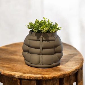 May include: A dark gray ceramic planter shaped like a puffer jacket with a small green plant inside. The planter is sitting on a wooden table.