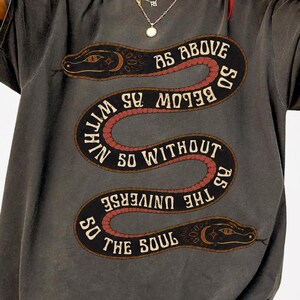 以下が含まれることがあります： チャコールグレーの半袖Tシャツで、蛇がうねる大きなグラフィックが描かれています。蛇の体には「AS ABOVE SO BELOW AS WITHIN SO WITHOUT AS THE UNIVERSE SO THE SOUL」という文字が記されています。デザインにはクリーム色と赤茶色の色調が使われています。