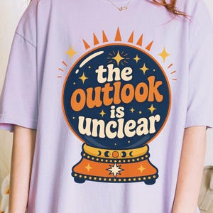 May include: A light purple short-sleeve t-shirt featuring a graphic of a dark blue crystal ball on a stand. The text "the outlook is unclear" is printed in orange and cream letters inside the ball, surrounded by small yellow star icons.