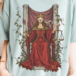May include: A light sage green t-shirt featuring a graphic print of a blindfolded figure in a red robe sitting on a stone throne. The figure holds a sword in one hand and a scale in the other, with vines climbing the pillars and the Roman numeral XI.