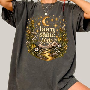 Może przedstawiać: Ciemnoszary t-shirt z nadrukiem graficznym przedstawiającym półksiężyc, gwiazdy, góry, krętą ścieżkę i kwiaty. Napis "born UNDER THE same Stars" znajduje się na środku wzoru, otoczony zielonymi liśćmi i żółtymi kwiatami.