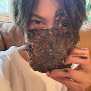 May include: A white t-shirt with the word "Pug" printed in black. A person is holding a rough, brown, textured object in front of their face. Their hands and face have dark smudges.