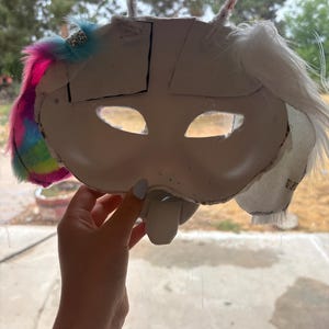 Puede incluir: Una máscara blanca con dos aberturas para los ojos y una pieza nasal prominente. El lado izquierdo está adornado con piel sintética multicolor y una cuenta plateada, mientras que el derecho presenta piel sintética blanca. Dos limpiapipas blancos sobresalen de la parte superior.