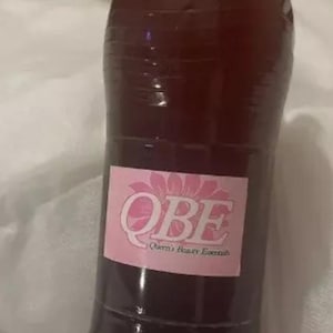 May include: A clear plastic bottle with a blue cap filled with a dark brown liquid. The bottle has a pink label with the text "QBE Queen's Beauty Essentials".
