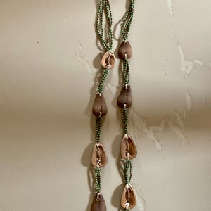 Può includere: Una lunga collana di perline verdi con baccelli di semi marroni che pendono da essa. La collana è composta da più fili di perline e ha un fiocco in fondo.