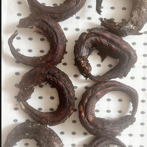 Puede incluir: Primer plano de ocho pescados secos dispuestos en círculo sobre una superficie blanca con lunares negros. Los pescados son de color marrón oscuro y tienen una textura ligeramente brillante.