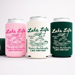 May include: Three beverage can coolers in pink, cream, and dark green. Each features the text "Lake Life" and a mermaid riding a fish, with "August 2026" and "Taylor's Bachelorette Lake Harmony" printed below.