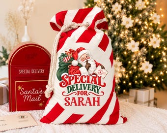 Pode incluir: Um saco de presente de tecido branco com listras diagonais vermelhas, apresentando um gráfico do Papai Noel segurando uma carta e um saco de presentes. O texto diz Special Delivery For Sarah. O saco tem um fecho de corda branca grossa.