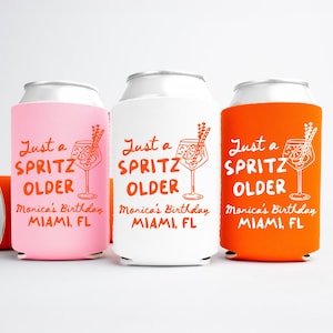 May include: Three beverage can coolers in pink, white, and orange, each with the text "Just a Spritz Older" and a cocktail illustration. The text "Monica's Birthday Miami, FL" is also printed on each cooler. One orange cooler is lying on its side.