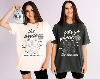 May include: A cream t-shirt with black text reading the bride and KATE'S BACHELORETTE, and a charcoal t-shirt with white text reading let's go ghouls and KATE'S BACHELORETTE. Both feature graphic illustrations of two ghosts, bats, and a disco ball.