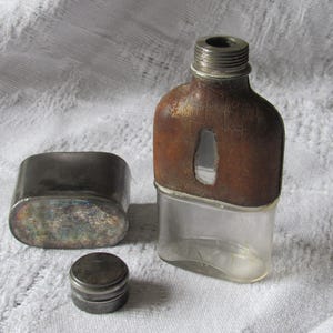 May include: An antique flask with a brown leather cover and a clear glass base. A small, round metal cap and a rectangular metal container are next to the flask. The flask has a threaded metal top.