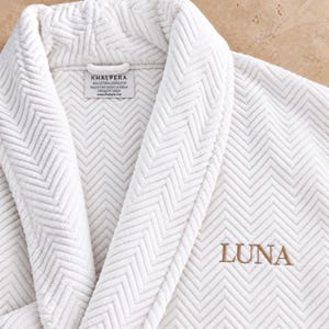 May include: A white bathrobe featuring a textured herringbone pattern and a shawl collar. The garment includes a label reading KHALPERA 100% COTTON ALGODON/COTON MADE IN TURKEY HECHO EN TURQUIA FABRIQUE EN TURQUIE and the name LUNA embroidered in gold.