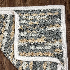 May include: A handmade crochet blanket with a shell stitch pattern. The blanket features a color scheme of gray, beige, and white, with a white border. The blanket is folded over, showing the texture and design.