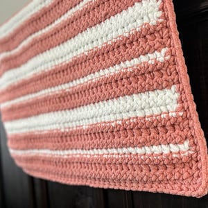 May include: A handmade, rectangular crochet blanket with alternating stripes of white and coral-pink. The blanket is made with a thick, textured yarn, and the stitches are clearly visible. The blanket is soft and cozy.