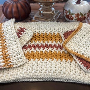 May include: Cream-coloured crocheted placemat with a central band of mustard yellow and burgundy stripes. Two matching square coasters are placed on either side. The placemat and coasters are part of a fall-themed home decor set.