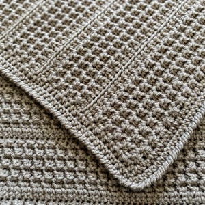 May include: Close-up of a handmade, gray crochet blanket. The blanket features a textured pattern with a waffle-like design and a ribbed border. The soft, neutral color and detailed stitching suggest a cozy, handcrafted item.