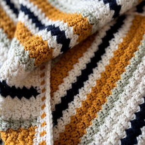 May include: Close-up of a handmade crochet blanket. The blanket features a repeating pattern of diagonal stripes in mustard yellow, navy blue, white, and pale green. The texture of the yarn is visible, creating a cozy and inviting aesthetic.