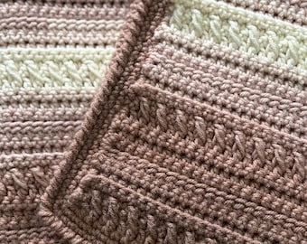 May include: Close-up of a crocheted blanket with a gradient of pink and white stripes. The blanket is made with a textured stitch and has a fringed edge.