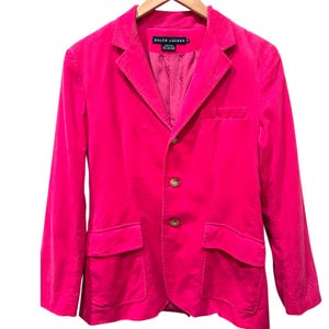 Pode incluir: Um blazer de veludo cotelê rosa brilhante com fecho de dois botões, dois bolsos frontais e um bolso no peito. O blazer tem lapelas entalhadas e está pendurado em um cabide de madeira. A etiqueta diz "RALPH LAUREN".