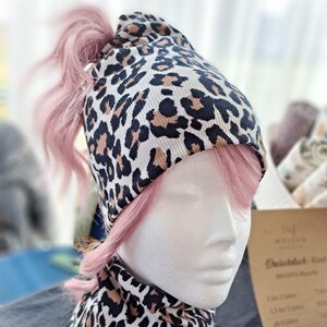 Könnte beinhalten: Ein Set aus Beanie-Mütze und Halstuch mit Leopardenmuster. Die Mütze hat eine Öffnung für einen Pferdeschwanz und wird mit einem passenden Halstuch kombiniert. Der Stoff hat einen cremefarbenen Hintergrund mit schwarzen und braunen Flecken. Das Set wird auf einem Schaufensterkopf mit rosa Haaren präsentiert.