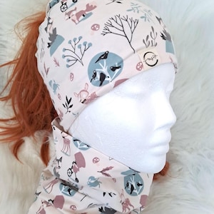 Könnte beinhalten: Cremefarbene Ponytail-Beanie und passendes Halstuch mit einem skurrilen Waldmuster mit Rehen, Vögeln und Bäumen in gedämpften Blau-, Rosa- und Grautönen. Die Beanie hat ein kleines Etikett mit einem Logo.