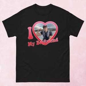 May include: Black t-shirt with a red heart-shaped graphic that says "I My Boyfriend." The heart contains a photo of a person flexing their arm. The shirt is a short-sleeved, casual style.