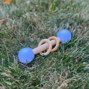 May include: A wooden baby rattle with two light blue golf balls on each end and three wooden rings in the middle. The rattle is lying in green grass.