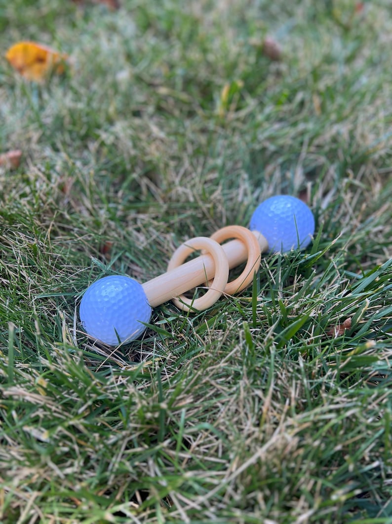 May include: A wooden baby rattle with two light blue golf balls on each end and three wooden rings in the middle. The rattle is lying in green grass.