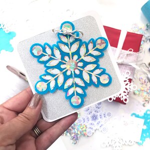 May include: A turquoise snowflake ornament with iridescent accents and a central gem, set on a silver glitter background. The ornament is tied with twine and sits on a square card. Other snowflake decorations are scattered around.