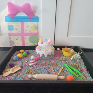 May include: A black sensory tray filled with colorful rice, sprinkles, and birthday-themed toys. There is a white cake with candles, a rolling pin, a scoop, a yellow bowl, and a gift box with a pink bow.