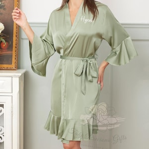 Puede incluir: Bata de satén verde salvia con mangas acampanadas y dobladillo con volantes. La bata tiene un cinturón a juego y el nombre "Hyeok" bordado en el pecho. Un regalo personalizado.
