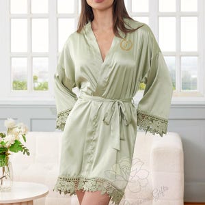 May include: Sage green satin robe with a matching tie belt and lace trim on the sleeves and hem. The robe features a gold embroidered initial on the chest. The robe is knee-length and has wide sleeves.