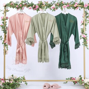 May include: Three satin robes in pink, sage green, and emerald green hang on a gold rack. Each robe has lace trim on the sleeves and hem, with a matching tie belt. The robes are personalized with names embroidered on the front.