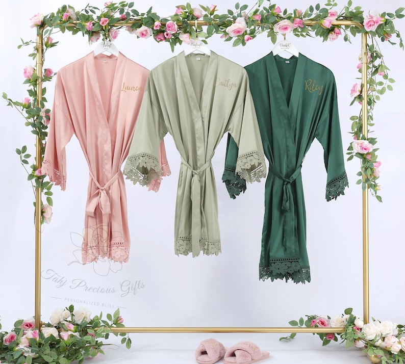 May include: Three satin robes in pink, sage green, and emerald green hang on a gold rack. Each robe has lace trim on the sleeves and hem, with a matching tie belt. The robes are personalized with names embroidered on the front.