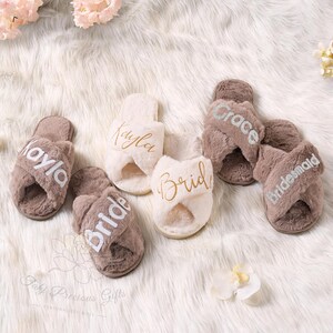 May include: A collection of plush, cross-band slippers in shades of brown and cream. Some slippers are personalized with names like "Kayla" and "Grace", and titles like "Bride" and "Bridesmaid" in elegant script. The slippers rest on a fluffy, white surface.