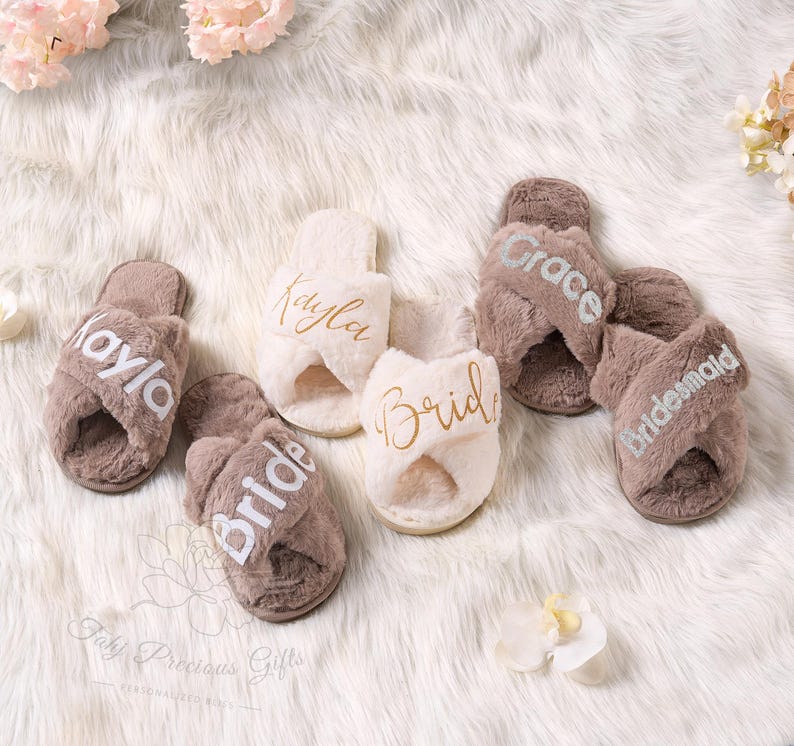May include: A collection of plush, cross-band slippers in shades of brown and cream. Some slippers are personalized with names like "Kayla" and "Grace", and titles like "Bride" and "Bridesmaid" in elegant script. The slippers rest on a fluffy, white surface.