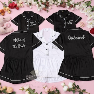 Pode incluir: Conjuntos de pijama de cetim preto e branco com debrum branco e texto personalizado. Os conjuntos incluem uma camisa com botões e calções. O texto inclui "Dion", "Lily", "Mother of the Bride", "Bridesmaid" e "Patsy". Apresentados numa superfície rosa com detalhes florais.