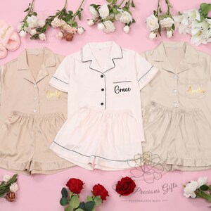 May include: Three sets of matching pajamas in beige and pink with black trim. Each set has a personalized name embroidered on the pocket: Emily, Grace, and Amelia. The pajamas are made of satin and have short sleeves and shorts.
