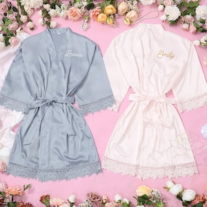 Peut inclure: Deux peignoirs en satin, un bleu clair et un rose pâle, sont présentés sur une surface rose entourée de fleurs. Le peignoir bleu clair porte le nom "Briana" brodé sur le devant. Le peignoir rose porte le nom "Emily". Les deux peignoirs ont une bordure en dentelle.