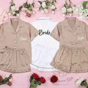 Puede incluir: Conjuntos de pijama de sat&eacute;n color beige y blanco, ribeteados con ribetes blancos, personalizados con nombres. Un conjunto blanco dice "Bride". Tambi&eacute;n se ven zapatillas rosas con nombres. Sobre una superficie rosa con flores.