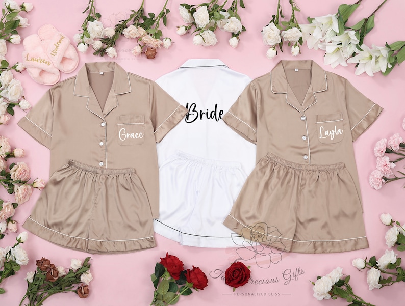 Puede incluir: Conjuntos de pijama de sat&eacute;n color beige y blanco, ribeteados con ribetes blancos, personalizados con nombres. Un conjunto blanco dice "Bride". Tambi&eacute;n se ven zapatillas rosas con nombres. Sobre una superficie rosa con flores.
