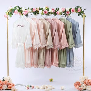 Könnte beinhalten: Eine Präsentation von Satin-Bademänteln in verschiedenen Pastellfarben, darunter Weiß, Rosa und Blau, die auf einem goldenen Gestell hängen. Der weiße Bademantel trägt den Aufdruck "Bride". Jeder Bademantel hat eine Spitzenausstattung. Das Gestell ist mit einem Blumengirlande verziert.