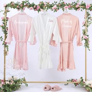 Puede incluir: Tres batas de seda en rosa y blanco cuelgan de un estante dorado decorado con flores. Las batas rosas est&aacute;n etiquetadas como "Bridesmaid" y "Maid of Honour". La bata blanca est&aacute; etiquetada como "Jade". Cada bata tiene un cintur&oacute;n a juego.