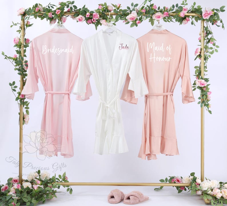 Puede incluir: Tres batas de seda en rosa y blanco cuelgan de un estante dorado decorado con flores. Las batas rosas est&aacute;n etiquetadas como "Bridesmaid" y "Maid of Honour". La bata blanca est&aacute; etiquetada como "Jade". Cada bata tiene un cintur&oacute;n a juego.