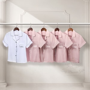 May include: Five personalised satin pyjamas with short sleeves and a button-down collar. The pyjamas are in pink and white with black trim. The names "Sophia", "Layla", "Olivia", "Amelia", and "Grace" are embroidered on the front of the pyjamas.