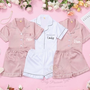 Può includere: Set di pigiami in raso rosa e bianco, con bordi a contrasto. Ogni set è personalizzato con un nome. I set sono disposti su una superficie rosa, circondati da fiori e una rivista.