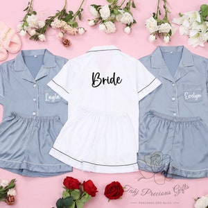 Puede incluir: Conjuntos de pijama a juego en azul claro y blanco, con ribetes blancos. Un conjunto blanco tiene la palabra "Bride" impresa en la espalda. Dos conjuntos azul claro tienen nombres impresos en la parte delantera. Los conjuntos incluyen camisa con botones y pantalones cortos. Zapatillas rosas y flores rodean los conjuntos.