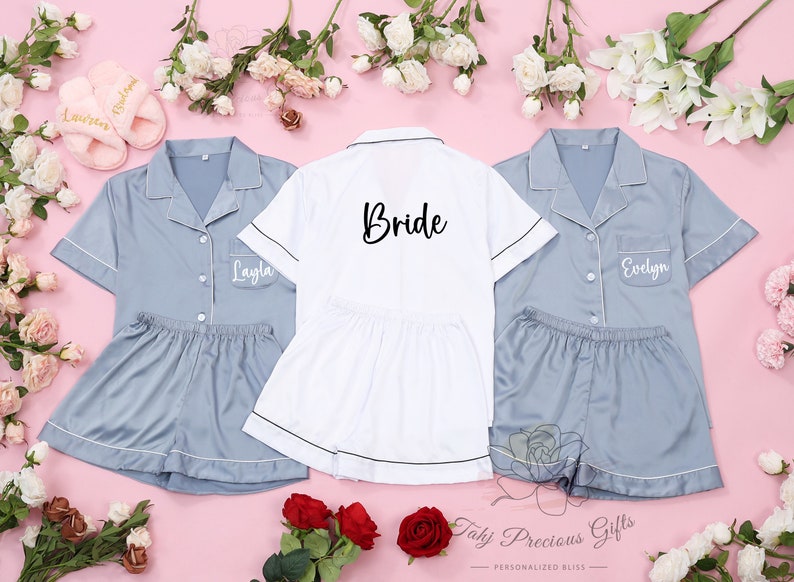 Puede incluir: Conjuntos de pijama a juego en azul claro y blanco, con ribetes blancos. Un conjunto blanco tiene la palabra "Bride" impresa en la espalda. Dos conjuntos azul claro tienen nombres impresos en la parte delantera. Los conjuntos incluyen camisa con botones y pantalones cortos. Zapatillas rosas y flores rodean los conjuntos.