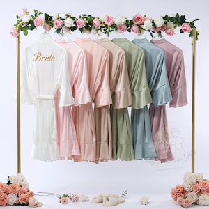 Op de afbeelding: Een collectie satijnen badjassen in verschillende pastelkleuren, waaronder wit met gouden "Bride" letters, roze, perzik, groen, blauw en lavendel, hangend aan een gouden rek. Het rek is versierd met een slinger van roze en witte rozen.